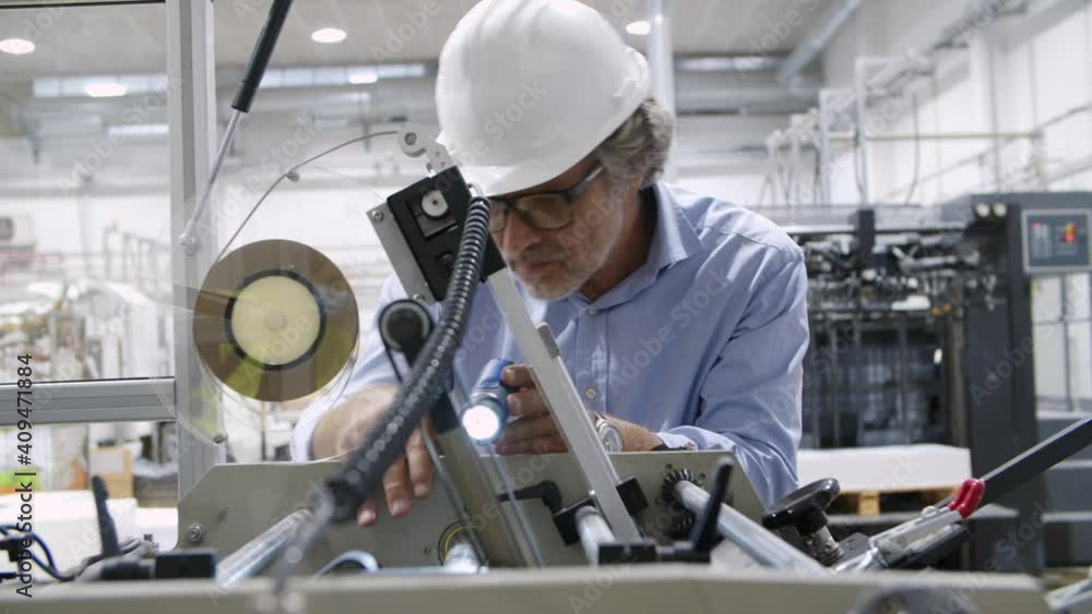 Focused old factory engineer inspecting machine with flashlight. Professional skilled worker in eyeglasses checking equipment on paper factory. Slider motion. Visual inspection and manufacture concept Focused old factory engineer inspecting machine with flashlight. Professional skilled worker in eyeglasses checking equipment on paper factory. Slider motion. Visual inspection and manufacture concept