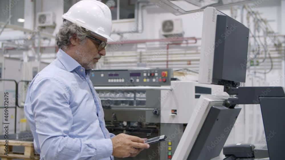 Older factory engineer testing industrial machine, pressing buttons at control board, monitoring process, using tablet. Production process or machinery concept Older factory engineer testing industrial machine, pressing buttons at control board, monitoring process, using tablet. Production process or machinery concept