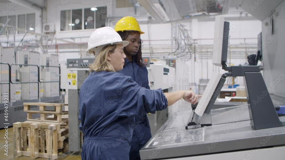 Middle aged female factory employee taking proficiency exam, explaining how to operate new machine to black mentor at control panel. Industry or machinery concept Middle aged female factory employee taking proficiency exam, explaining how to operate new machine to black mentor at control panel. Industry or machinery concept