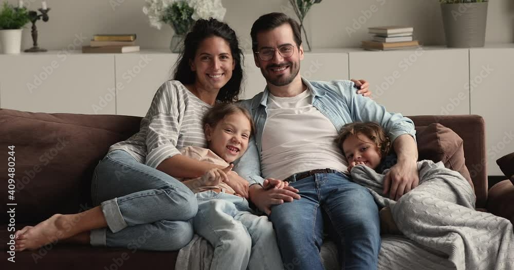 Portrait of happy lovely family with little kids sit on couch smile look at camera. Young parents rest on sofa hug preschool children spend weekend at modern home. Upbringing, happy parenthood concept Portrait of happy lovely family with little kids sit on couch smile look at camera. Young parents rest on sofa hug preschool children spend weekend at modern home. Upbringing, happy parenthood concept