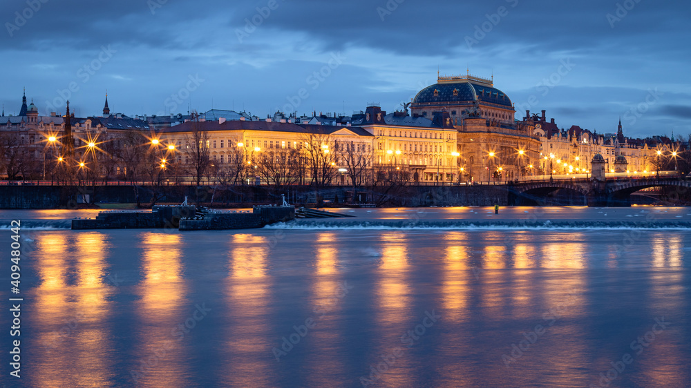 View of illuminated historic houses  and National Theater along Vltava river waterfront in Prague, Czech Republic at twilight