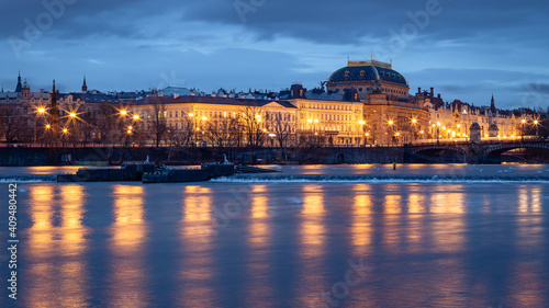 View of illuminated historic houses and National Theater along Vltava river waterfront in Prague, Czech Republic at twilight