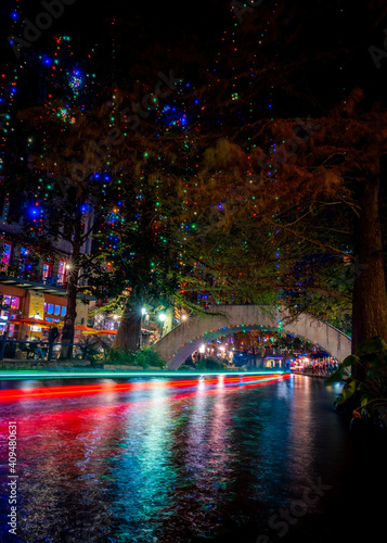 The River Walk in San Antonio, Texas at night with Christmas lights and riverboat neon light trails. Riverwalk tourist destination with arched bridge, restaurants, and shops