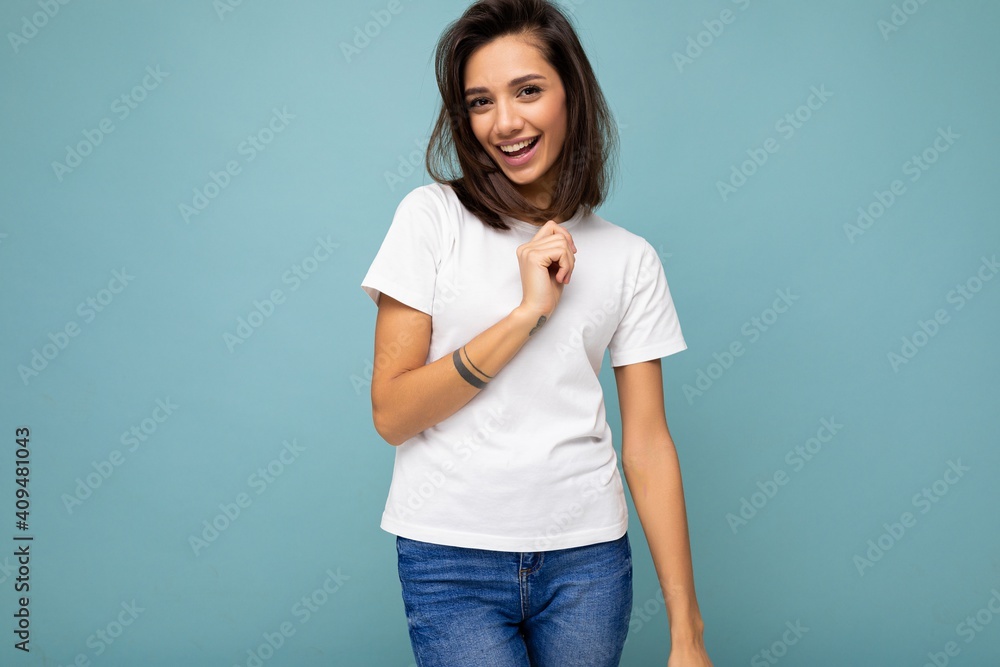 Portrait of positive cheerful fashionable smiling young brunette woman in casual white t-shirt for mockup isolated on blue background with copy space Portrait of positive cheerful fashionable smiling young brunette woman in casual white t-shirt for mockup isolated on blue background with copy space