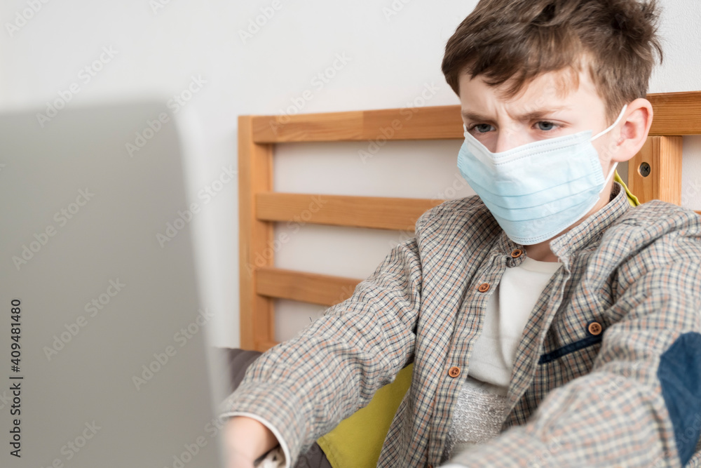Boy medical mask is watching school lesson laptop, sitting bed on pillows. Quarantine in world