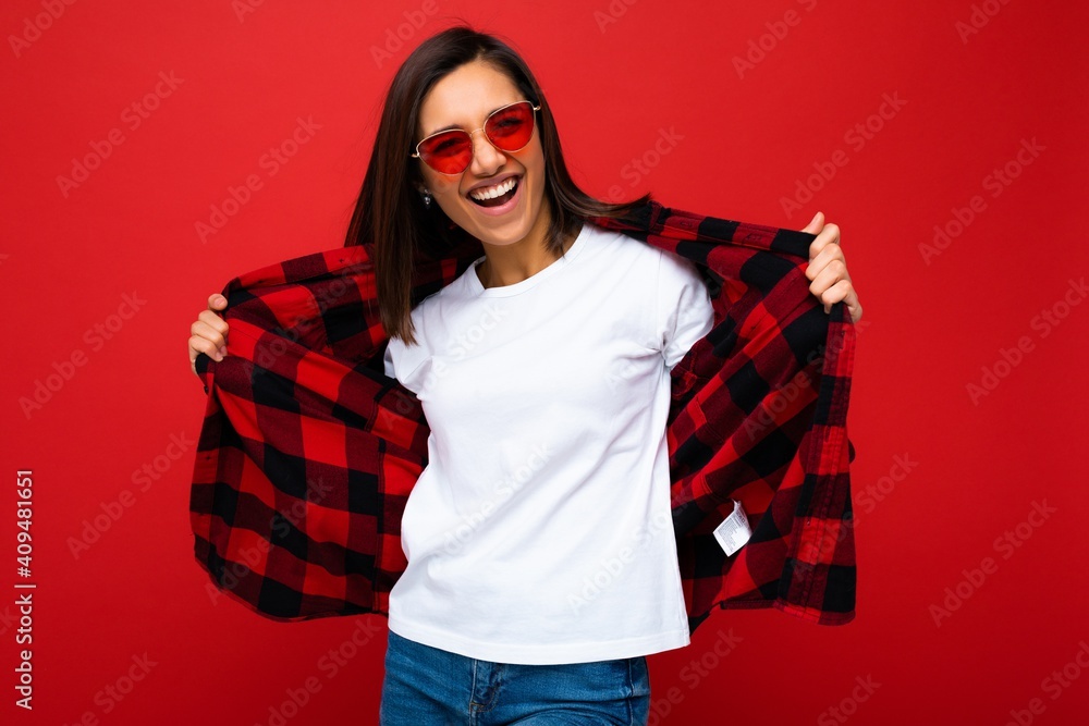 Positive happy smiling young brunette woman in casual white t-shirt for mockup, stylish red check shirt and trendy red sunglasses isolated on red background with copy space Positive happy smiling young brunette woman in casual white t-shirt for mockup, stylish red check shirt and trendy red sunglasses isolated on red background with copy space
