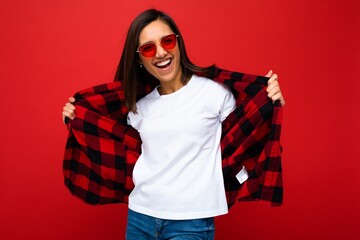 Positive happy smiling young brunette woman in casual white t-shirt for mockup, stylish red check shirt and trendy red sunglasses isolated on red background with copy space