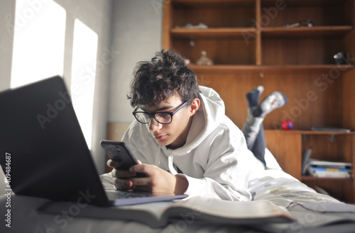 young boy studies lying on the bed using computer and smartphone Fototapeta