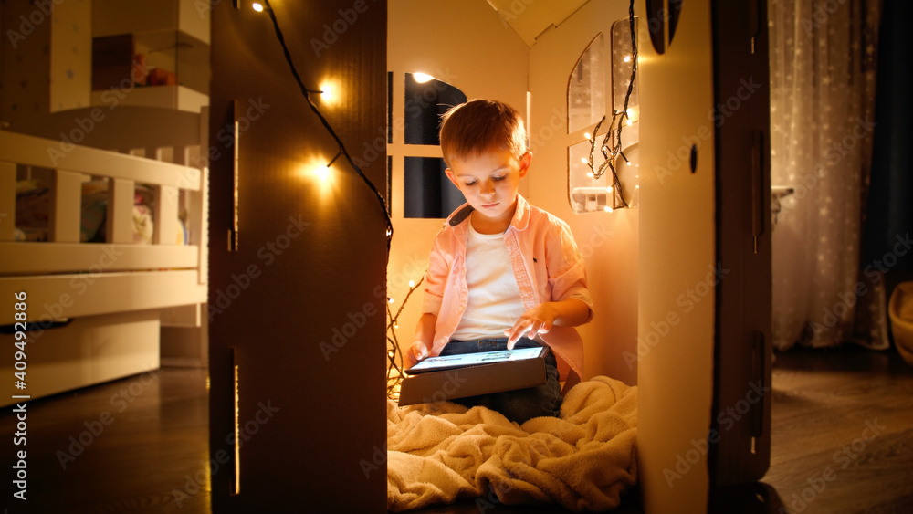 Little smart boy sitting in toy cardboard house and using tablet computer. Concept of child education and studying at night Little smart boy sitting in toy cardboard house and using tablet computer. Concept of child education and studying at night