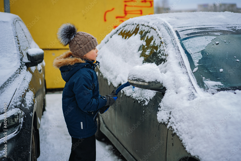 cute little caucasian boy wearing  knit hat and hooded jacket brushing snow from a car on winter day . Image with selective focus