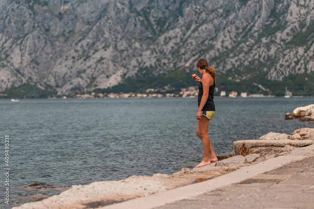 A young athletic woman stands on the shore and eats an apple in the summer .Kotor bay (Boka Kotorska), Montenegro, Europe