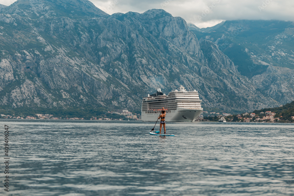 A young athletic woman stands on a SUP stand up paddle board in the Bay of Kotor with a large cruise ship in the background .Kotor bay (Boka Kotorska), Montenegro, Europe
