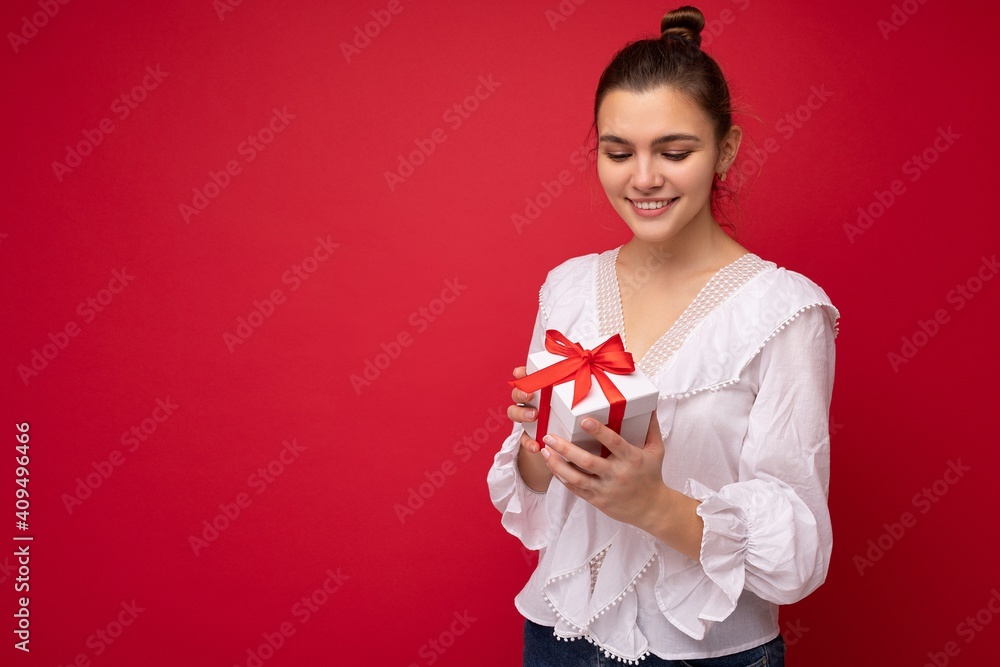 Shot of charming happy smiling young brunette woman isolated over red background wall wearing white blouse holding white gift box with red ribbon and looking down Shot of charming happy smiling young brunette woman isolated over red background wall wearing white blouse holding white gift box with red ribbon and looking down