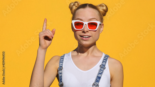 Portrait happy teen blonde girl in stylish sunglasses confidently raises her index finger up with delight looks at camera on yellow background in summer. Emotions of people. Positive mood