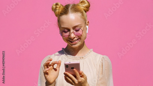 Young happy blonde girl teen blogger makes selfie on smartphone in wireless headphones  in pink sunglasses and smiles slow motion on pink background summer. Emotions of people. Relax. Youth. Monotone
