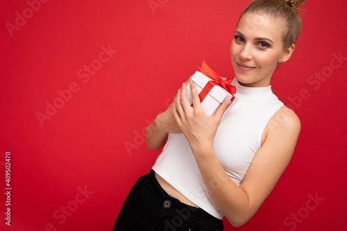 Fascinating sexy positive smiling young blonde woman isolated over red background wall wearing white top holding gift box and looking at camera