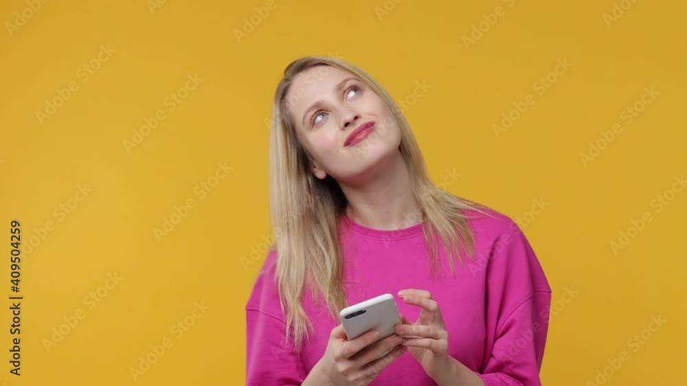 Pensive smiling blonde young woman 20s years old in casual pink t-shirt posing isolated on yellow background studio. People lifestyle concept. Using mobile cell phone typing browsing looking aside up Pensive smiling blonde young woman 20s years old in casual pink t-shirt posing isolated on yellow background studio. People lifestyle concept. Using mobile cell phone typing browsing looking aside up