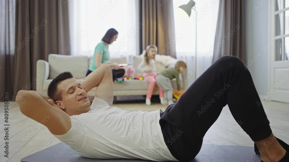 Active lifestyle. Sporty man training at home, doing crunches while family playing on background, slow motion Active lifestyle. Sporty man training at home, doing crunches while family playing on background, slow motion