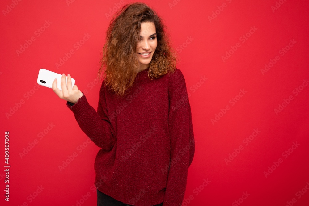 Naklejka premium Photo of pretty smiling happy young woman with curly hair wearing dark red sweater isolated on red background holding mobile phone looking to the side and having fun
