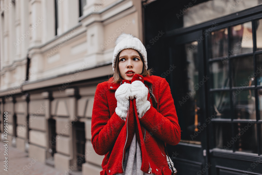 Girl in snow-white knitted hat and mittens is shaking from cold, wrapping herself around in red coat Girl in snow-white knitted hat and mittens is shaking from cold, wrapping herself around in red coat