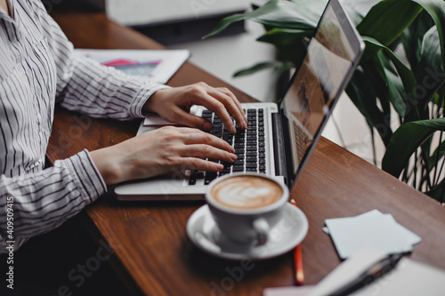 Woman works in laptop while sitting in cozy office at table. Shot of cup of coffee  sheets of paper and pen on wooden table