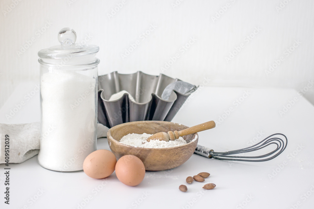 Homemade baking ingredients and accessories. A jar of sugar, a cup of flour, eggs, a whisk, a cake pan, a napkin, nuts on a white table. Home hobbies authentic cooking concept, close-up, copy space Homemade baking ingredients and accessories. A jar of sugar, a cup of flour, eggs, a whisk, a cake pan, a napkin, nuts on a white table. Home hobbies authentic cooking concept, close-up, copy space