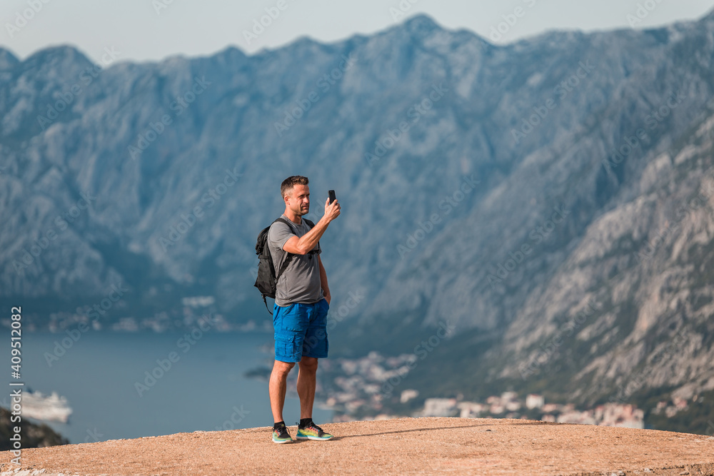 Young athletic traveler with a backpack making selfie with smartphone on the top of the mountain at Kotor bay (Boka Kotorska), Montenegro, Europe