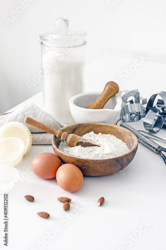 Homemade baking ingredients and accessories. A jar of sugar, a cup of flour, eggs, a whisk, a cake pan, a napkin, nuts on a white table. Home hobbies authentic cooking concept, close-up, copy space