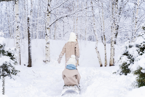 Little beautiful stylish girl sledding her sister. Winter hat, woolen coat, blue scarf, snood. Kids walking, playing in forest, park among trees covered with snow.Country house yard.Fashionable image