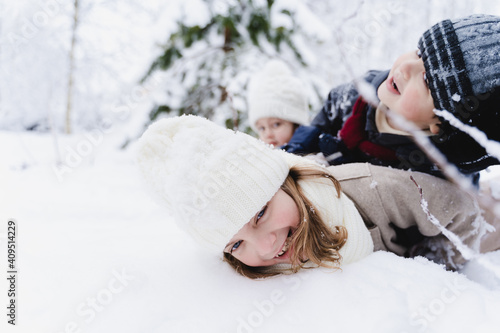 Family fun,winter kids activity in snowy clothes with mom.Children lay in heap on top of each other.Happy cute boys,girls.Walking in beautiful forest,park.Snowy trees.Hat,coat,scarf.Fashionable image