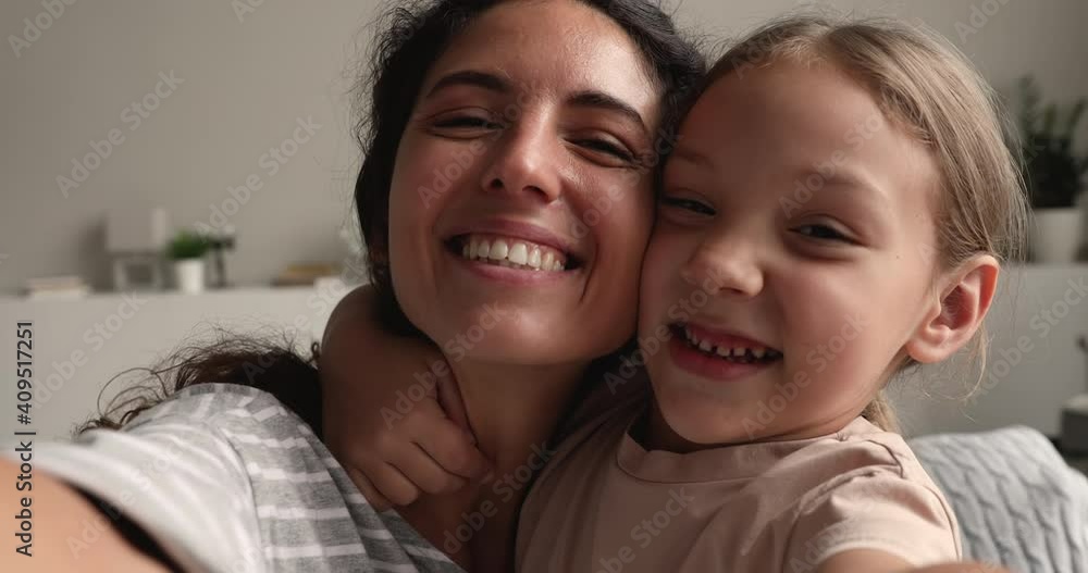 Young mom her little daughter hugging hold device smile look at camera record tender moment, webcam view. Close up faces loving mother and kid touch cheeks making video call feels happy and overjoyed Young mom her little daughter hugging hold device smile look at camera record tender moment, webcam view. Close up faces loving mother and kid touch cheeks making video call feels happy and overjoyed