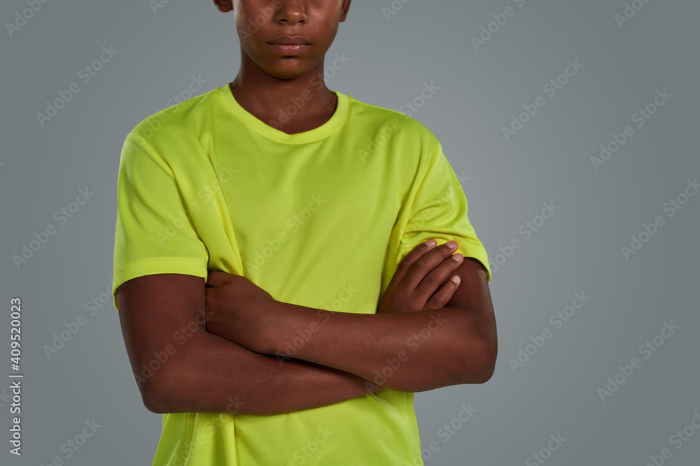 Teenage african boy wearing neon t shirt keeping arms crossed while posing isolated over grey background in studio Teenage african boy wearing neon t shirt keeping arms crossed while posing isolated over grey background in studio