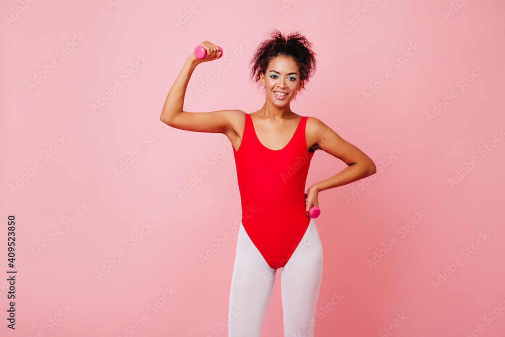 Glad african woman in aerobics form using dumbbells. Studio shot of winsome black girl training on pink background.