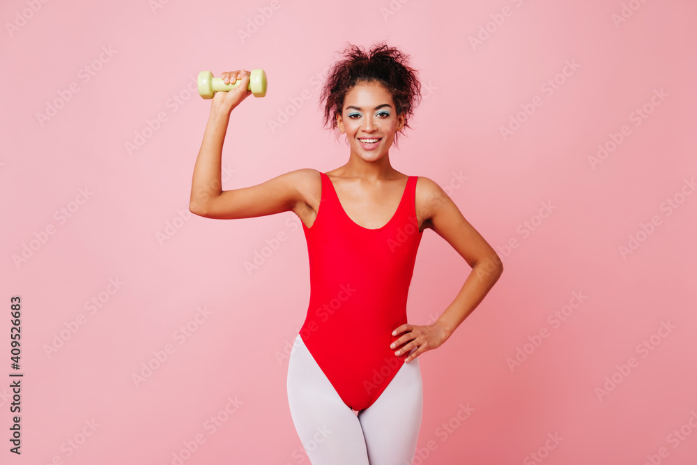 Fascinating woman using yellow dumbbell. Studio shot of refined african girl in aerobics bodysuit smiling on pink background. Fascinating woman using yellow dumbbell. Studio shot of refined african girl in aerobics bodysuit smiling on pink background.