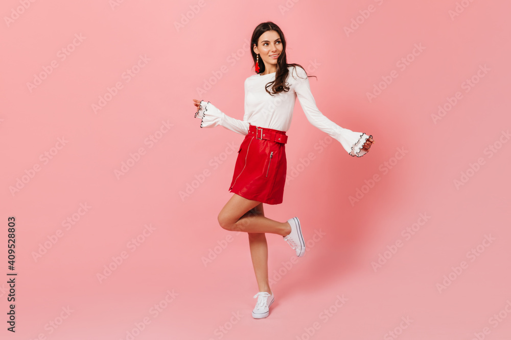 Full-length portrait of young dark-haired girl in leather red skirt and light blouse dancing on pink background