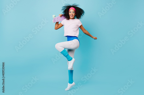 Carefree fitness woman dancing with sincere smile. Studio shot of slim african girl jumping with skateboard.