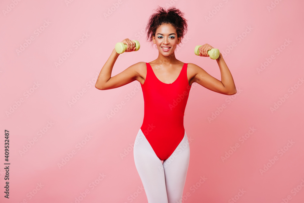 Enthusiastic female model in aerobics bodysuit smiling on pink background. Studio shot of slim african girl training with dumbbells. Enthusiastic female model in aerobics bodysuit smiling on pink background. Studio shot of slim african girl training with dumbbells.