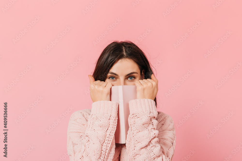 Joyful dark-haired girl covers her face with her pink notebook. Woman in knitted sweater looks into camera Joyful dark-haired girl covers her face with her pink notebook. Woman in knitted sweater looks into camera