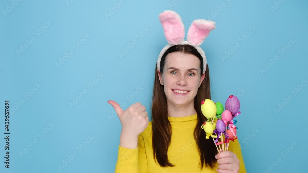 Young excited woman girl 20s in pink bunny fluffy ears, say hey you look pointing fingers hands on copy space aside workspace, holding small eggs, isolated on blue background studio. Easter concept Young excited woman girl 20s in pink bunny fluffy ears, say hey you look pointing fingers hands on copy space aside workspace, holding small eggs, isolated on blue background studio. Easter concept