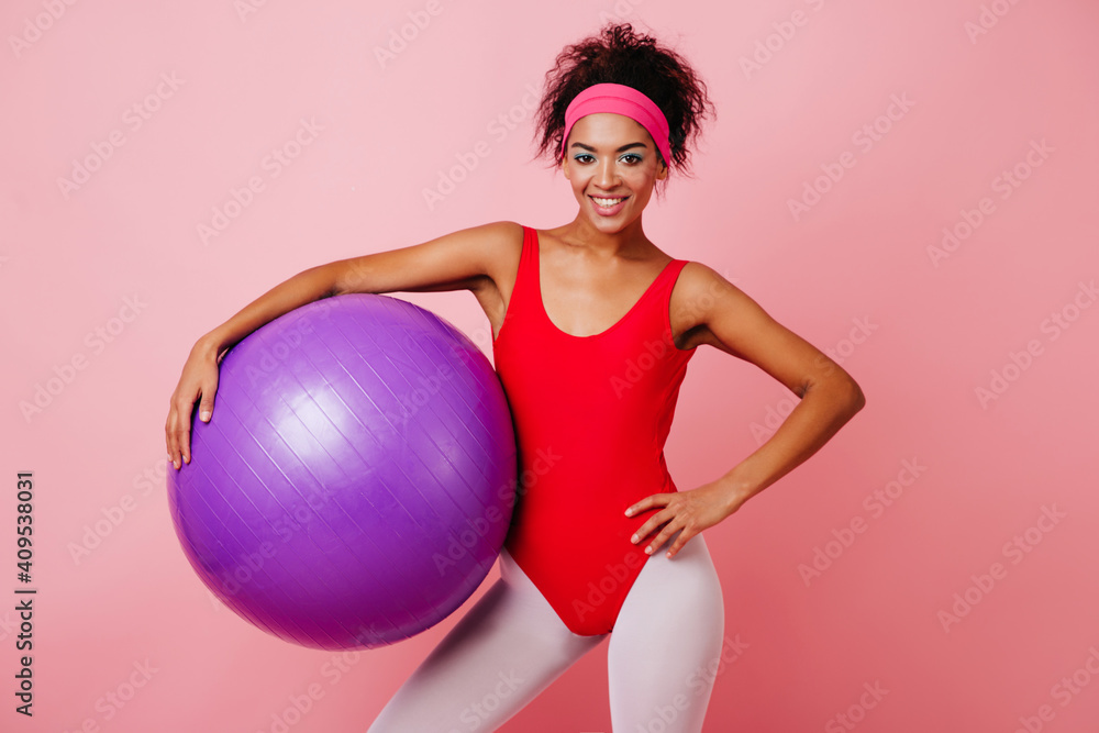 Charismatic girl with fitness ball laughing to camera. Confident african woman in vintage sportswear posing on pink background. Charismatic girl with fitness ball laughing to camera. Confident african woman in vintage sportswear posing on pink background.