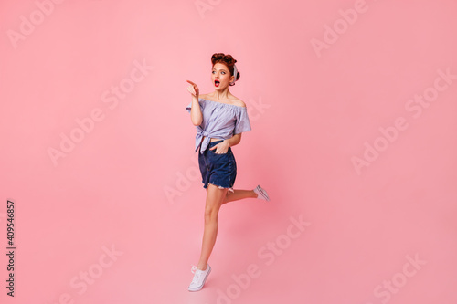 Surprised woman in denim shorts pointing with finger. Amazed pinup lady jumping on pink background.