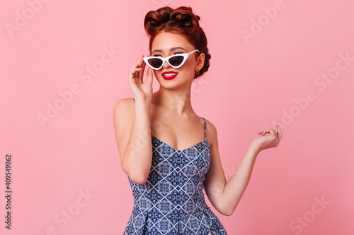 Romantic ginger girl touching sunglasses. Studio shot of pinup woman in blue dress.