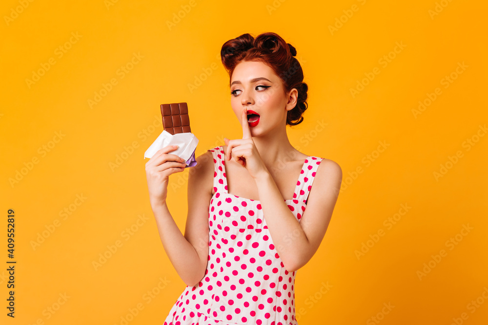 Sensual pinup woman eating chocolate on yellow background. Ginger girl in polka-dot dress touching lips with finger. Sensual pinup woman eating chocolate on yellow background. Ginger girl in polka-dot dress touching lips with finger.