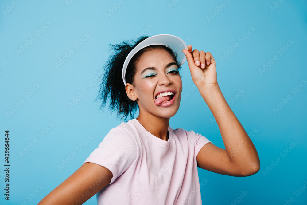 Playful fitness girl posing with tongue out. Studio shot of happy african lady touching tennis hat. Playful fitness girl posing with tongue out. Studio shot of happy african lady touching tennis hat.