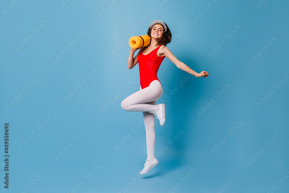 Girl in tight spots suit happily jumps on blue background. 80s woman carries her yellow fitness mat Girl in tight spots suit happily jumps on blue background. 80s woman carries her yellow fitness mat