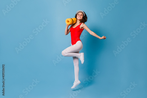Fototapeta Naklejka Na Ścianę i Meble -  Girl in tight spots suit happily jumps on blue background. 80s woman carries her yellow fitness mat