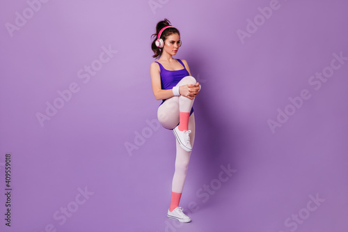 Fototapeta Naklejka Na Ścianę i Meble -  Woman in headphones listens to music and does warm-up before fitness workout on purple background