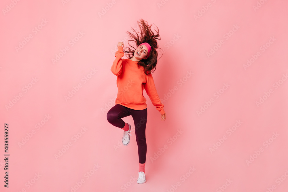 Lady in bright sweatshirt and brown leggings dances against pink background. Fitness trainer enjoying workout - obrazy, fototapety, plakaty 