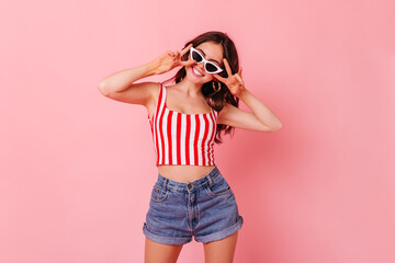tylish dark-haired woman in summer shorts and top smiles and shows peace sign on pink background