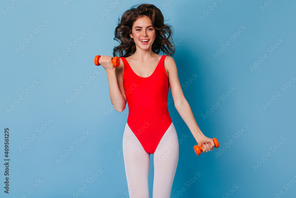 Active brunette girl in sports suit with smile jumping on blue background. Woman in leggings holds small dumbbells Active brunette girl in sports suit with smile jumping on blue background. Woman in leggings holds small dumbbells